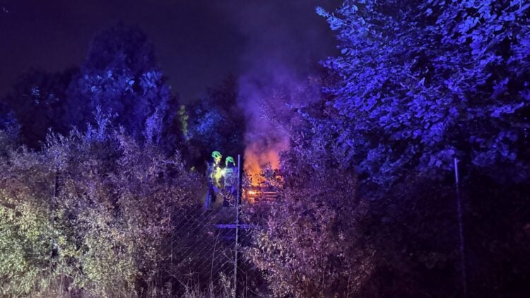 Zwei Feuerwehrleute mit Helmen hinter einem Maschendrahtzaun, daneben brennende Holzpaletten zwischen Büschen bei Nacht