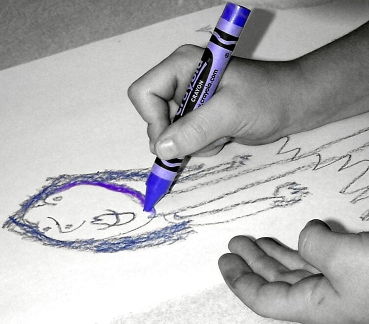 Kinderhand malt mit einem lila Wachsmalstift ein Gesicht auf Papier