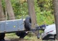 Beschädigtes weißes Auto an einem Baum, davor ein Anhänger, Polizisten untersuchen die Unfallstelle