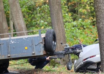 Beschädigtes weißes Auto an einem Baum, davor ein Anhänger, Polizisten untersuchen die Unfallstelle