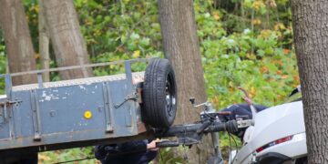 Beschädigtes weißes Auto an einem Baum, davor ein Anhänger, Polizisten untersuchen die Unfallstelle