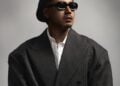 Rapper Yangpapi mit Baskenmütze, Sonnenbrille und dunklem Anzug vor grauem Hintergrund