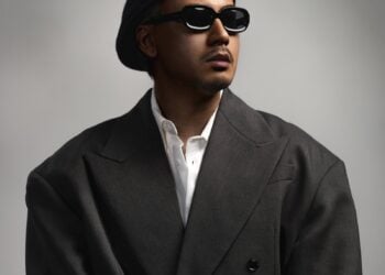 Rapper Yangpapi mit Baskenmütze, Sonnenbrille und dunklem Anzug vor grauem Hintergrund