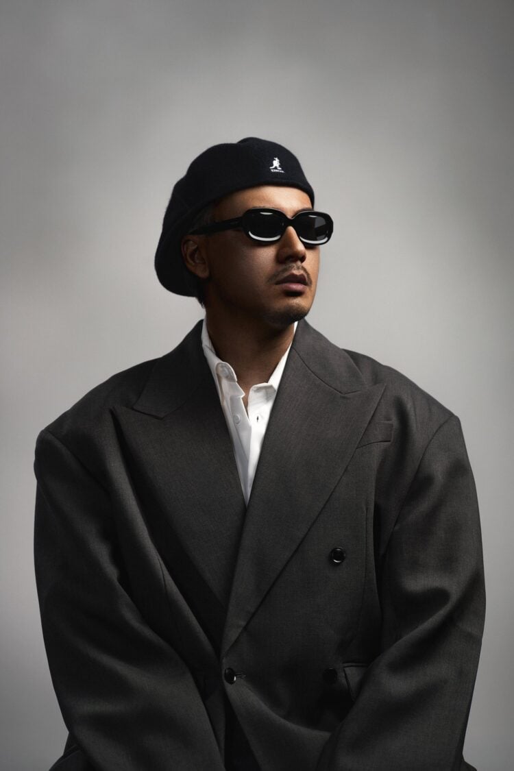 Rapper Yangpapi mit Baskenmütze, Sonnenbrille und dunklem Anzug vor grauem Hintergrund