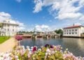 Fluss mit Brücke, links ein weißes Eckgebäude, rechts ein Industriebau, im Vordergrund Blumen