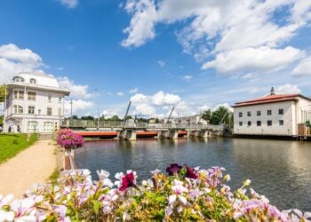 Fluss mit Brücke, links ein weißes Eckgebäude, rechts ein Industriebau, im Vordergrund Blumen