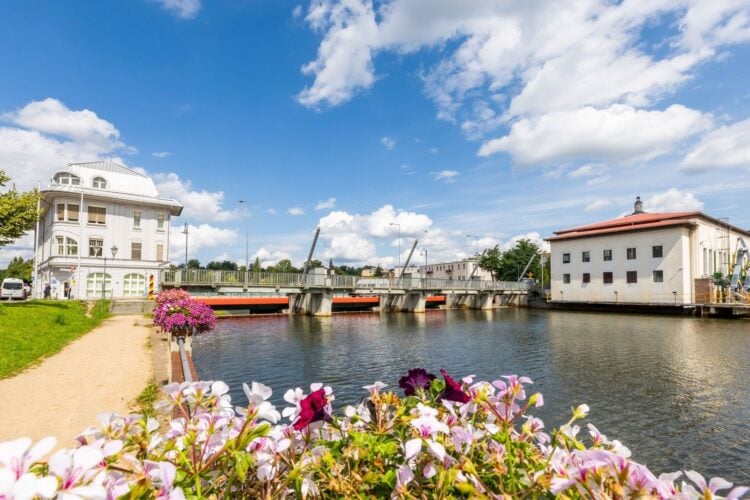 Fluss mit Brücke, links ein weißes Eckgebäude, rechts ein Industriebau, im Vordergrund Blumen