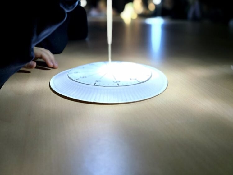Eine selbst gebastelte Sonnenuhr aus Pappe liegt im Lichtkegel auf einem Tisch, daneben die Hand eines Kindes