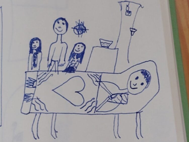 Kinderzeichnung mit Stift: Eine Familie steht an einem Krankenbett, auf der Bettdecke ist ein großes Herz gemalt