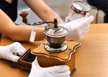 Hände in weißen Baumwollhandschuhen untersuchen eine alte Kaffeemühle und eine Porzellantasse auf einem Tisch