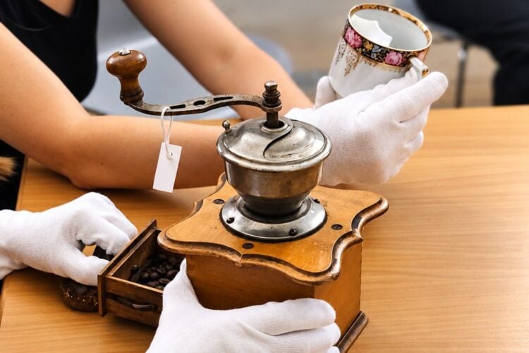 Hände in weißen Baumwollhandschuhen untersuchen eine alte Kaffeemühle und eine Porzellantasse auf einem Tisch