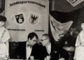 Schwarz-Weiß-Foto, zwei Männer an einem Tisch vor einer Tafel mit den Wappen von Neunkirchen und Lübben, daneben zwei Frauen in Tracht