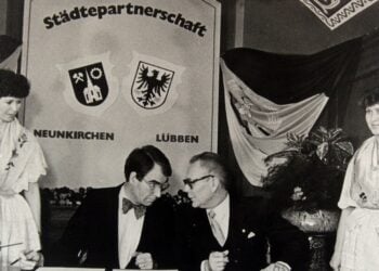 Schwarz-Weiß-Foto, zwei Männer an einem Tisch vor einer Tafel mit den Wappen von Neunkirchen und Lübben, daneben zwei Frauen in Tracht
