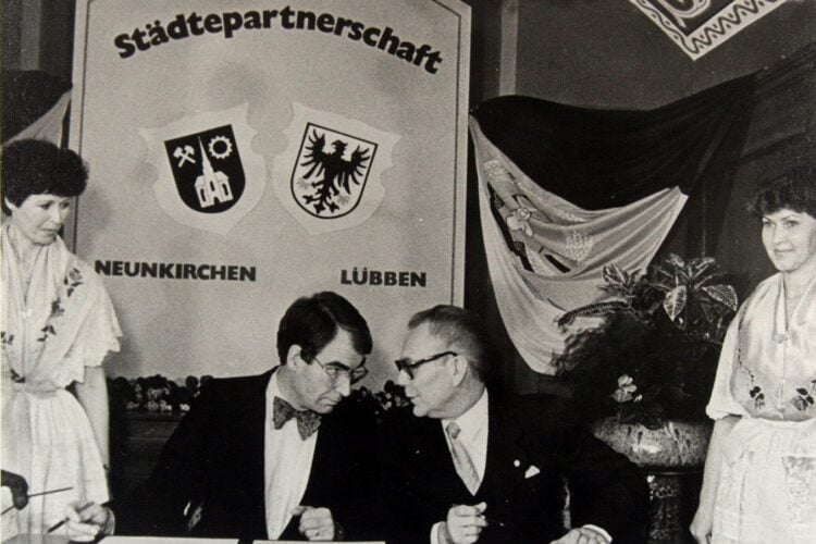 Schwarz-Weiß-Foto, zwei Männer an einem Tisch vor einer Tafel mit den Wappen von Neunkirchen und Lübben, daneben zwei Frauen in Tracht