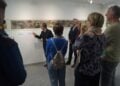 Ein Künstler zeigt in der Galerie auf ein langes Bild an der Wand, Besucher hören zu