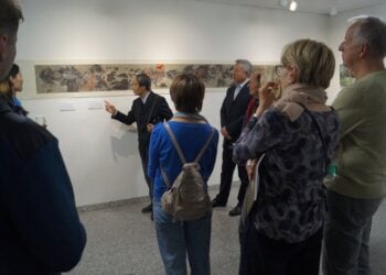 Ein Künstler zeigt in der Galerie auf ein langes Bild an der Wand, Besucher hören zu