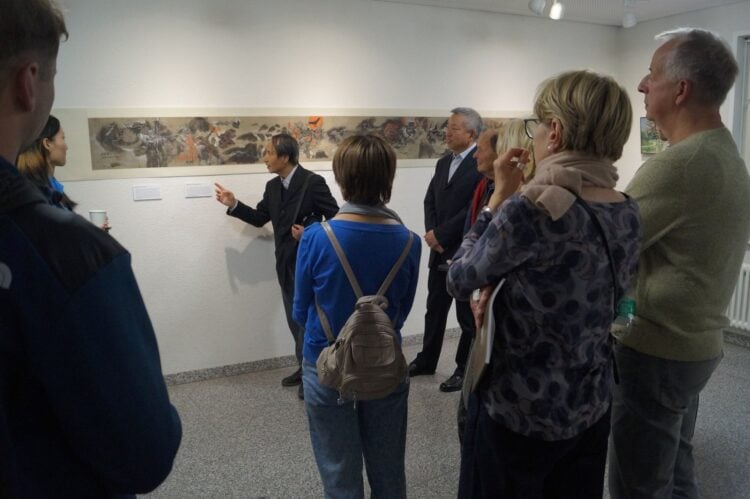 Ein Künstler zeigt in der Galerie auf ein langes Bild an der Wand, Besucher hören zu