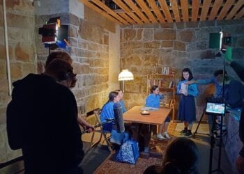 Jugendliche in blauen Kostümen spielen an einem Tisch in einem Raum mit Sandsteinwänden, im Vordergrund Tonangel, Scheinwerfer und Kameramonitor