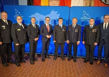 Acht Männer in Feuerwehruniform und Anzug stehen nebeneinander vor einer blauen Wand mit dem Logo des Landes Brandenburg