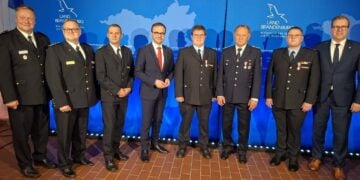 Acht Männer in Feuerwehruniform und Anzug stehen nebeneinander vor einer blauen Wand mit dem Logo des Landes Brandenburg