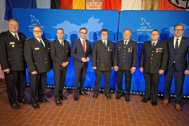 Acht Männer in Feuerwehruniform und Anzug stehen nebeneinander vor einer blauen Wand mit dem Logo des Landes Brandenburg