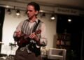 Schauspieler Karsten Zinser in Hemd und gestreifter Weste spielt Ukulele, im Hintergrund ein Schild mit der Aufschrift Brecht Bar