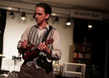 Schauspieler Karsten Zinser in Hemd und gestreifter Weste spielt Ukulele, im Hintergrund ein Schild mit der Aufschrift Brecht Bar
