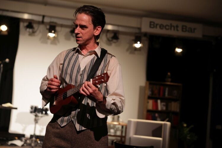 Schauspieler Karsten Zinser in Hemd und gestreifter Weste spielt Ukulele, im Hintergrund ein Schild mit der Aufschrift Brecht Bar