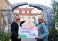 Zwei Männer halten vor einem mit grauer Folie verhüllten Torbogen ein Schild der KWG und der neuen Bühne Senftenberg