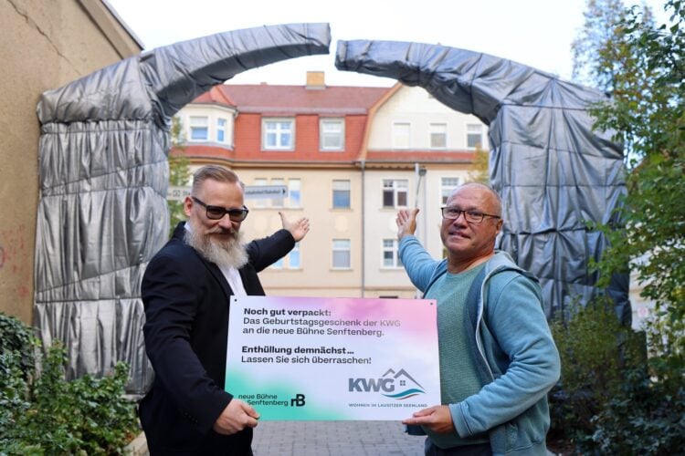 Zwei Männer halten vor einem mit grauer Folie verhüllten Torbogen ein Schild der KWG und der neuen Bühne Senftenberg