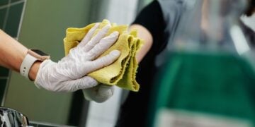 Hand in weißem Einweghandschuh poliert mit gelbem Tuch einen Spiegel über einem Waschbecken