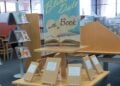 In Packpapier eingeschlagene Bücher mit Beschreibungszetteln auf einem Bibliothekstisch, dahinter ein Schild Blind Date with a Book