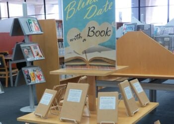 In Packpapier eingeschlagene Bücher mit Beschreibungszetteln auf einem Bibliothekstisch, dahinter ein Schild Blind Date with a Book
