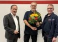 Drei Männer vor einer weißen Wand mit rotem Streifen, der mittlere hält einen Blumenstrauß und schüttelt dem rechten die Hand
