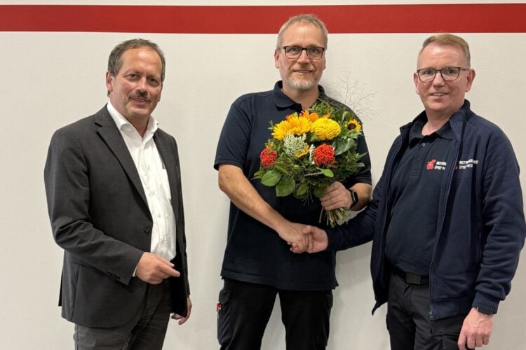 Drei Männer vor einer weißen Wand mit rotem Streifen, der mittlere hält einen Blumenstrauß und schüttelt dem rechten die Hand
