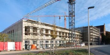 Rohbau eines dreigeschossigen Schulgebäudes mit Gerüst und Baukränen an einer Straße, daneben ein historischer Klinkerbau