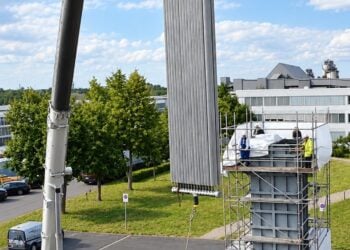 Ein Autokran hebt eine hohe, gerippte Heizfläche über einen eingerüsteten Speicherbehälter, auf dem Gerüst stehen Monteure
