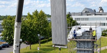 Ein Autokran hebt eine hohe, gerippte Heizfläche über einen eingerüsteten Speicherbehälter, auf dem Gerüst stehen Monteure