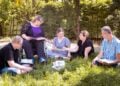 Menschen sitzen mit Arbeitsheften im Kreis auf einer Wiese, eine Frau im Rollstuhl