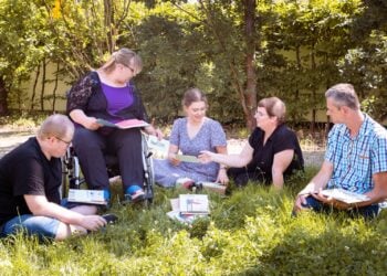 Menschen sitzen mit Arbeitsheften im Kreis auf einer Wiese, eine Frau im Rollstuhl