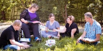 Menschen sitzen mit Arbeitsheften im Kreis auf einer Wiese, eine Frau im Rollstuhl