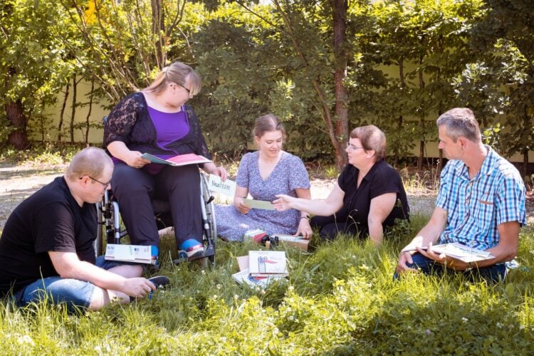 Menschen sitzen mit Arbeitsheften im Kreis auf einer Wiese, eine Frau im Rollstuhl