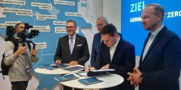 Vier Männer an einem Stehtisch auf einem Messestand, einer unterschreibt ein Dokument, dahinter eine Karte des Rheinischen Reviers