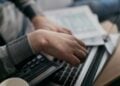 Hände rechnen mit Taschenrechner neben Laptop und Unterlagen auf einem Schreibtisch