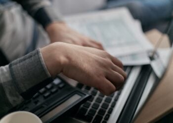 Hände rechnen mit Taschenrechner neben Laptop und Unterlagen auf einem Schreibtisch