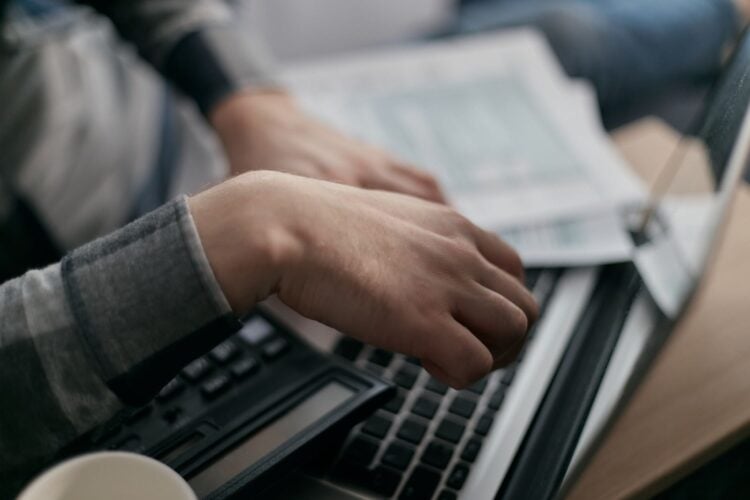 Hände rechnen mit Taschenrechner neben Laptop und Unterlagen auf einem Schreibtisch