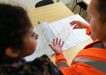 Zwei Personen besprechen einen ausgedruckten Lageplan auf einem Bürotisch, einer davon in orangefarbener Warnjacke