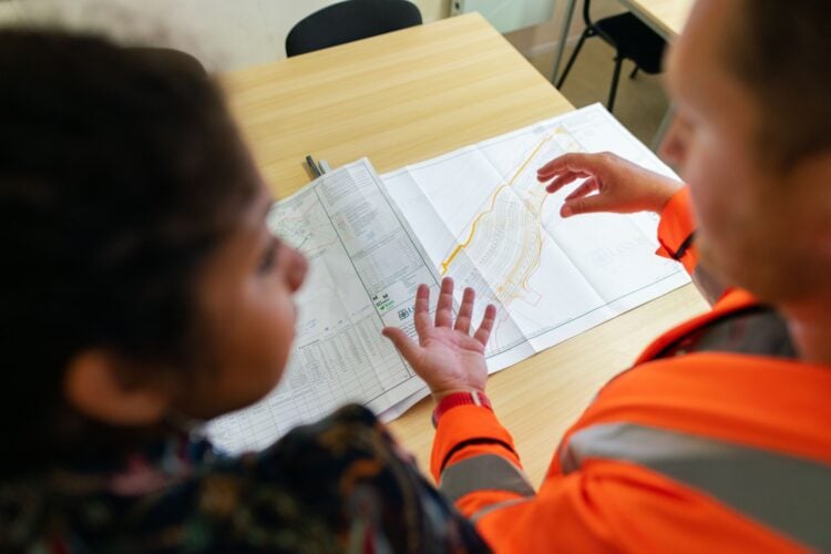 Zwei Personen besprechen einen ausgedruckten Lageplan auf einem Bürotisch, einer davon in orangefarbener Warnjacke