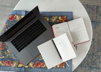 Aufgeklappter Laptop neben zwei offenen Notizbüchern und einem Stift auf gemustertem Stoff