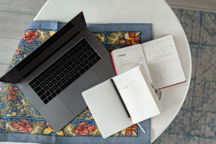 Aufgeklappter Laptop neben zwei offenen Notizbüchern und einem Stift auf gemustertem Stoff